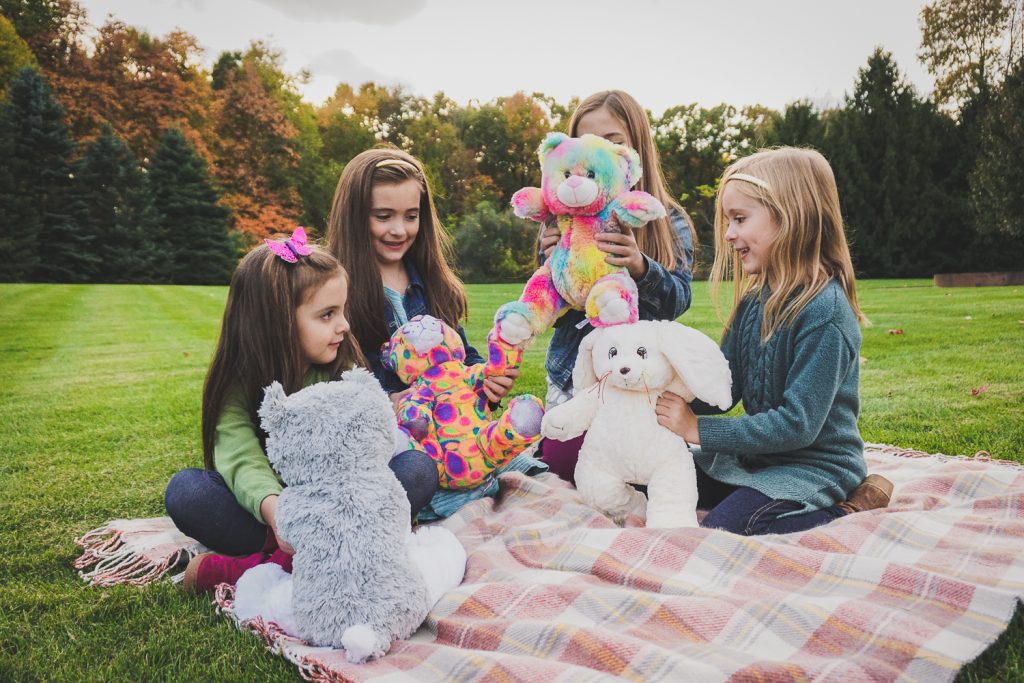 Four children playing with plush animals on a pink picnic blanket on grass. Click for an express order form.
